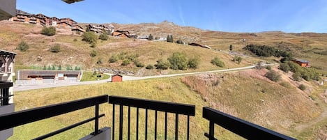 A balcony with a view of a hillside, a building with a sloped roof, and a clear sky.