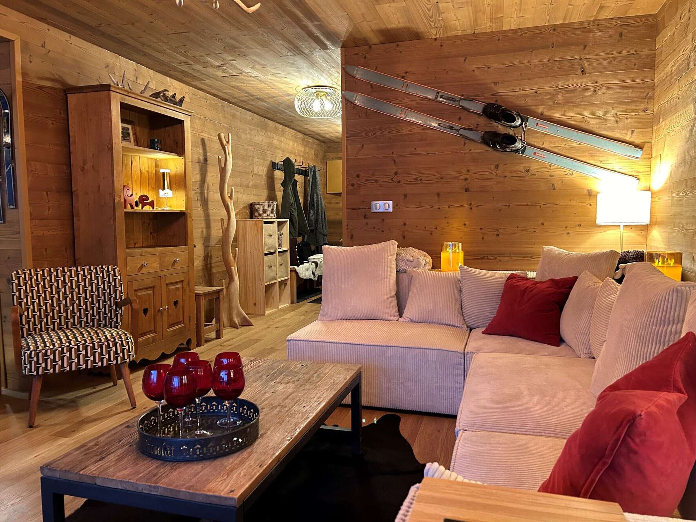 A cozy cabin interior with a wooden ceiling, a sofa, a table with wine glasses, and skis hanging on the wall.