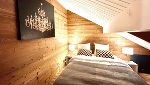 A bedroom with wooden walls, a sloped ceiling, a chandelier, a bed with checkered pillows, and bedside lamps.