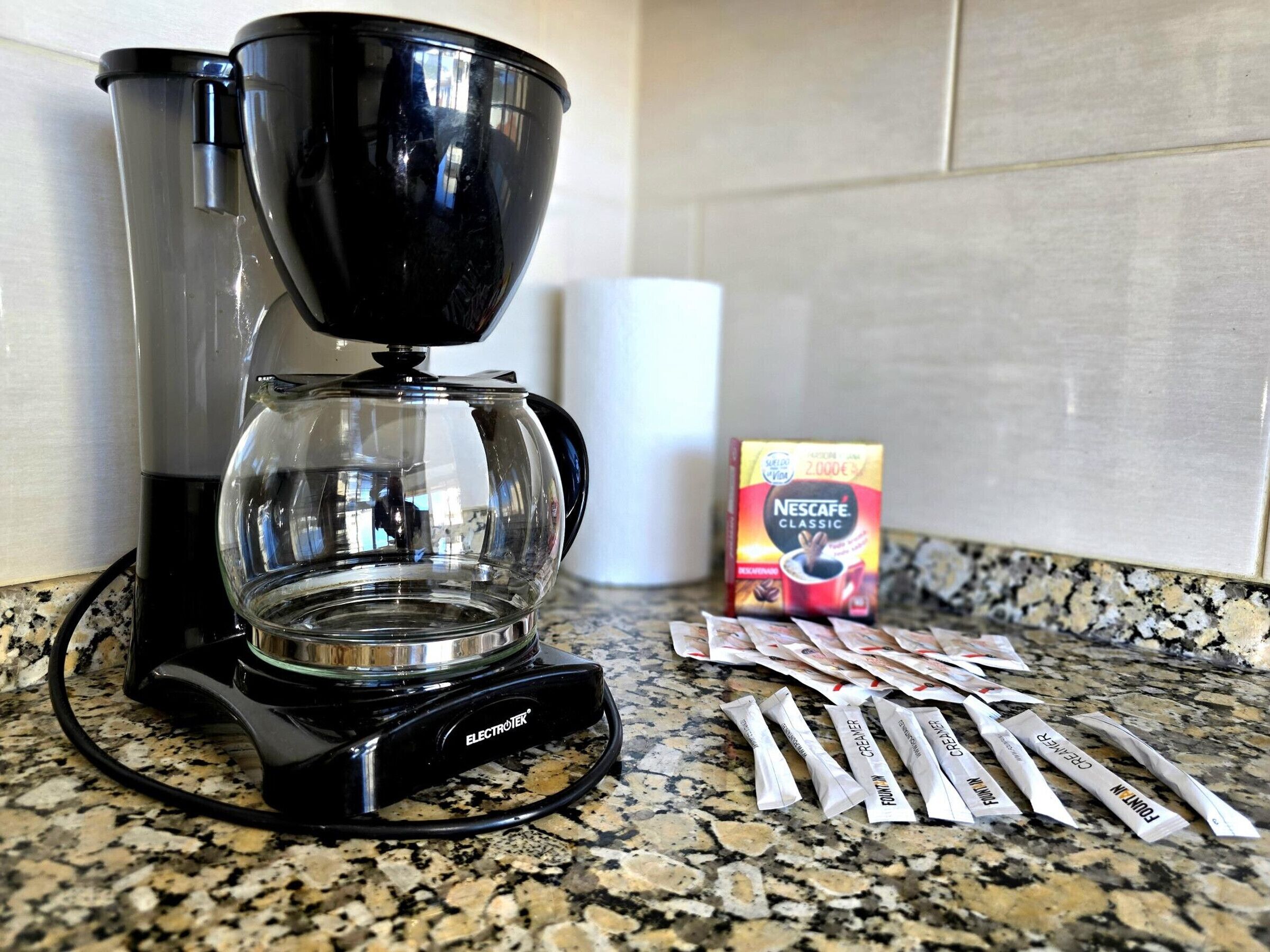 Une machine à café avec une verseuse en verre, une boîte de dosettes de café Nescafe Classic et plusieurs dosettes de café sur un comptoir en granit.
