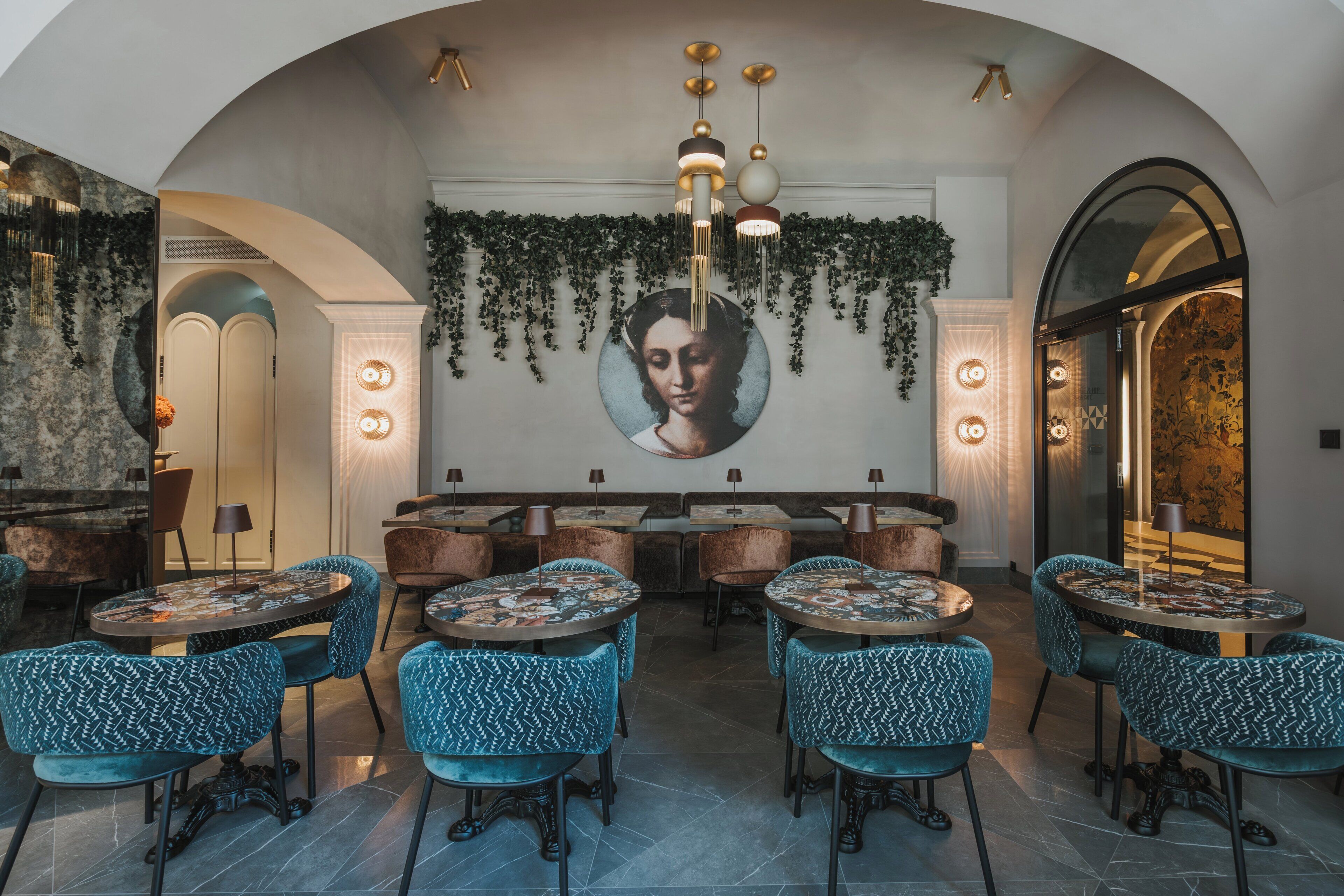 A restaurant with round tables and blue patterned chairs, a mural on the wall, and hanging plants.