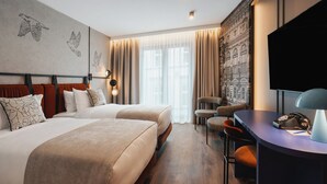 Una habitación de hotel con dos camas, un escritorio con lámpara, una silla y un ventanal con cortinas.