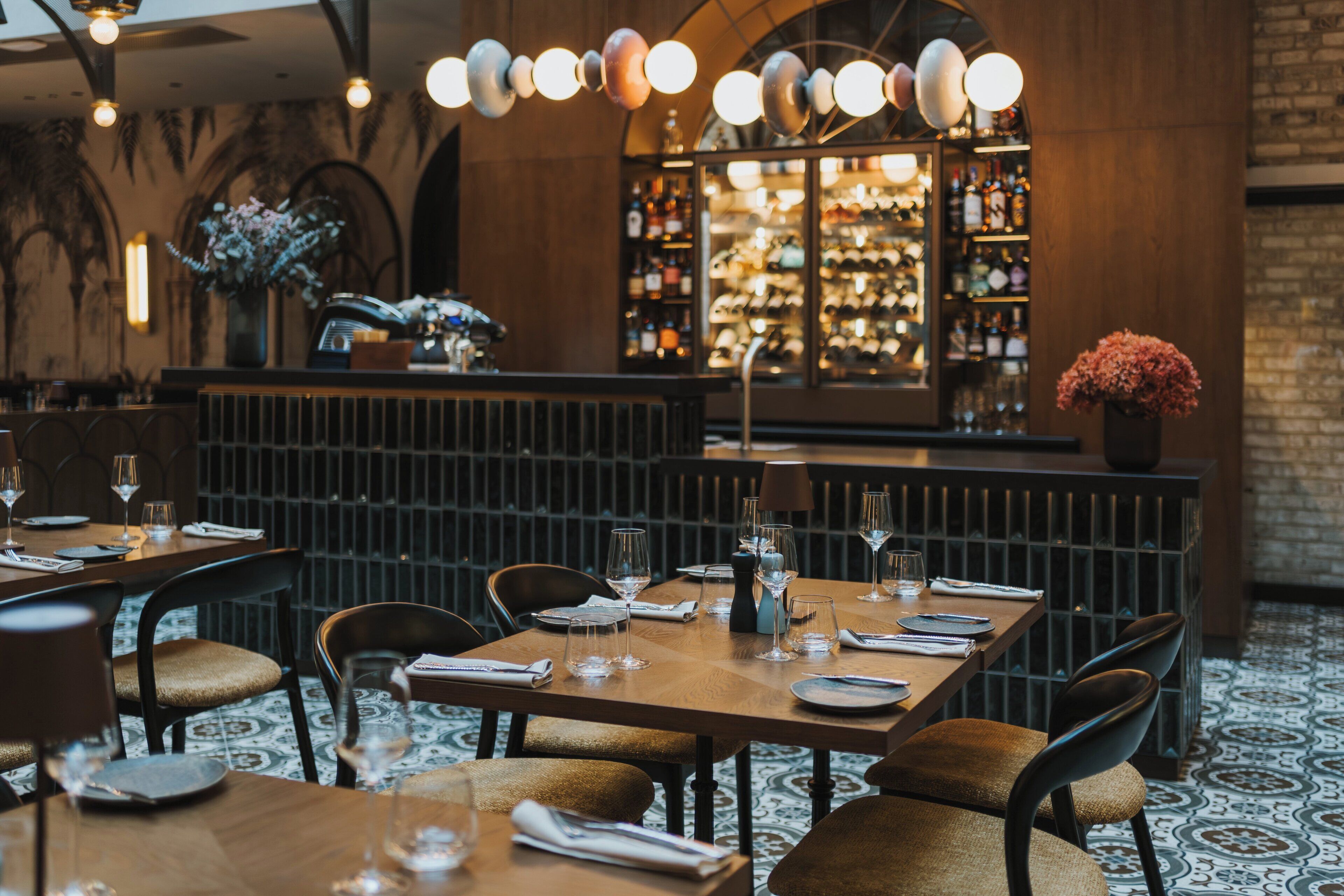 Restaurante com mesas e cadeiras de madeira, área de bar com garrafas e piso estampado.