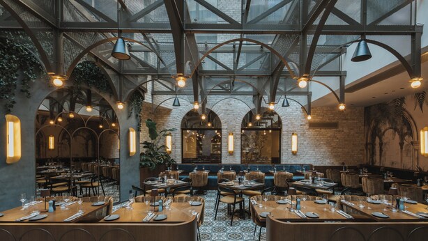 Un restaurante con techos altos, paredes de ladrillo visto y un suelo estampado. Las mesas están dispuestas con vajilla y velas.