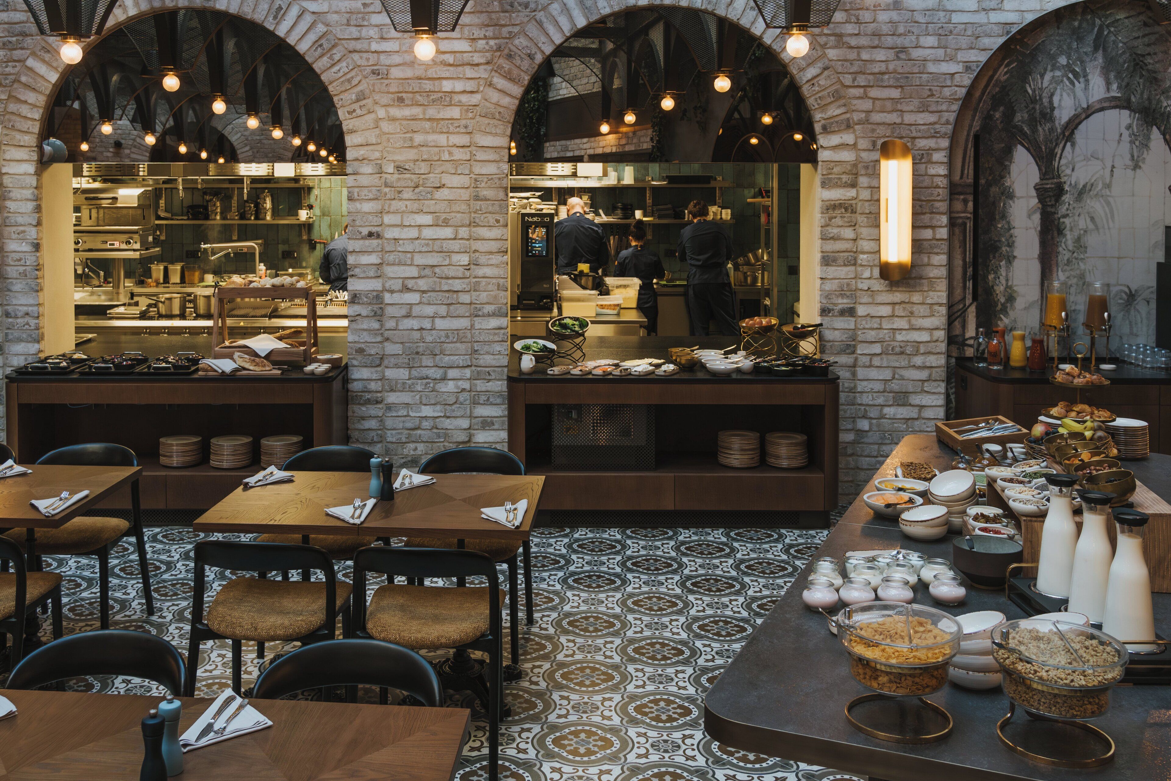 Un restaurante con paredes de ladrillo, techos abovedados y piso con diseño. Las mesas están dispuestas con sillas, y se observa una zona de bufé.