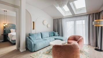 Ein modernes Wohnzimmer mit einem blauen Sofa, einem rosa Sessel und einem weißen Couchtisch. Ein großes Fenster mit Vorhängen und ein Oberlicht sind vorhanden.
