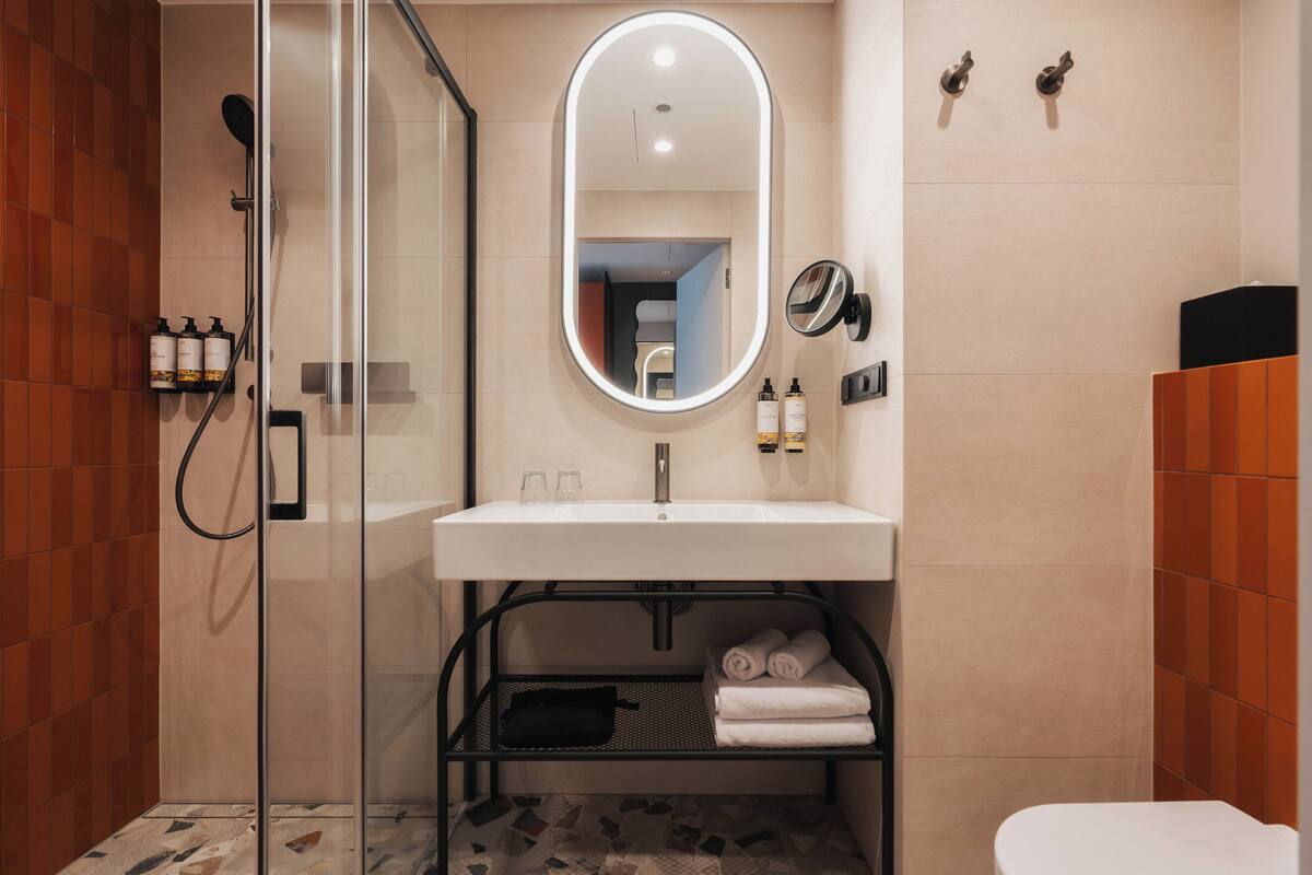 Un baño moderno con ducha de vidrio, un lavamanos blanco con espejo y una repisa con toallas.