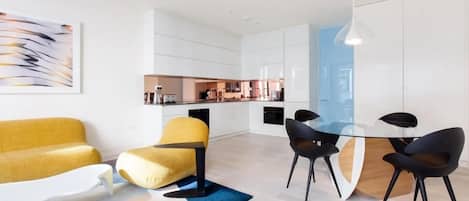 A modern living room with a yellow sofa, a glass dining table with black chairs, and a kitchen in the background.