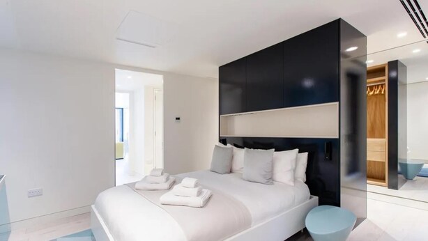 A modern bedroom with a large bed, a dark headboard, and a light-colored round ottoman. - Central London Charm One Bedroom Apartment (London)