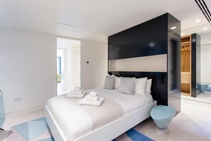 Une chambre à coucher moderne avec un grand lit, une tête de lit sombre et un pouf rond de couleur claire.