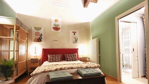 Un dormitori amb sostre de fusta, un llit amb almoades estampades, una taula de nit i una planta en test, i una porta que dona a una altra habitació.