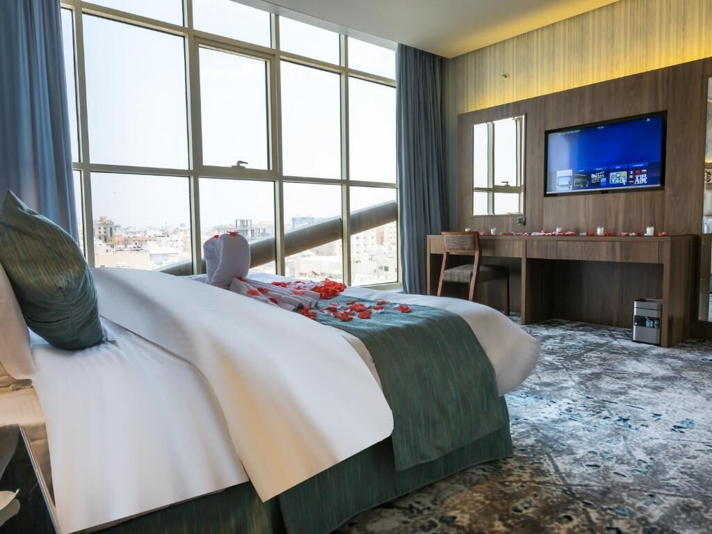 Une chambre d’hôtel avec un grand lit, un bureau, une télévision et une vue sur la ville grâce à de grandes fenêtres.