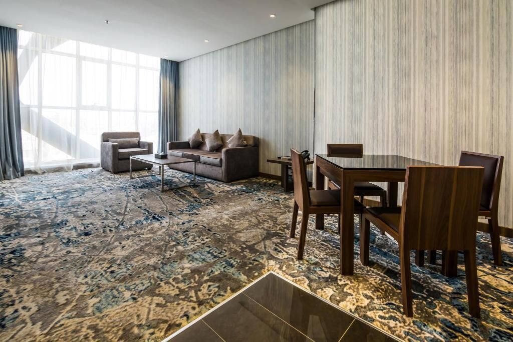 A modern hotel room with a large window, a sofa, a dining table with chairs, and a patterned carpet.