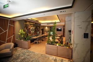 Una hall moderna di un hotel con pavimento in legno, un'area salotto centrale con tavoli e sedie, e piante decorative.
