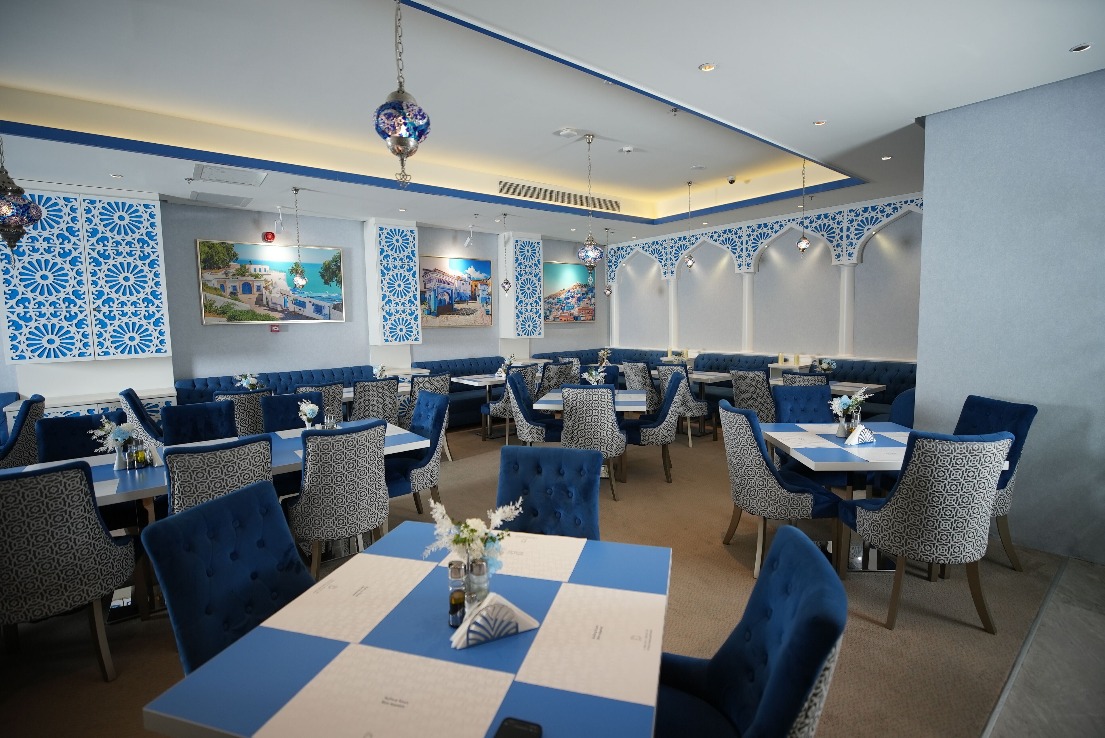 A restaurant with blue and white decor, featuring patterned chairs, tables with menus, and hanging decorative lights.