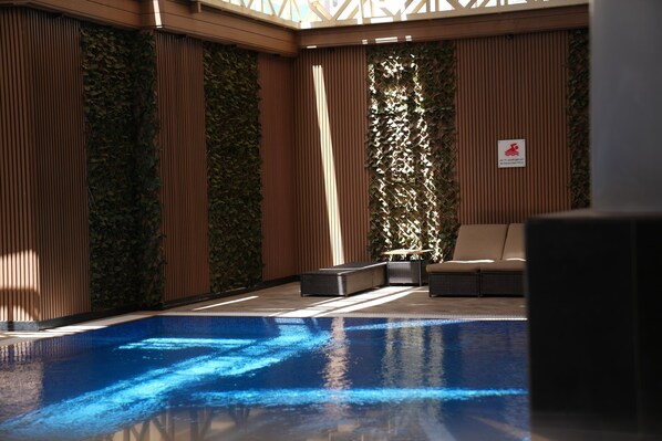 Un área interior de piscina moderna con una piscina azul cristalina, paneles de madera y una zona de estar con cojines.
