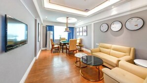 A modern living room with a sofa, coffee table, and TV. There are clocks on the wall and a dining area in the background. - Junfu Hotel (Guangzhou Panyu Square Metro Station) (Guangzhou)