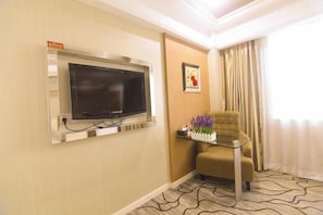 A hotel room with a mounted television, a small table with a vase of flowers, a comfortable armchair, and a patterned carpet. - Junfu Hotel (Guangzhou Panyu Square Metro Station) (Guangzhou)