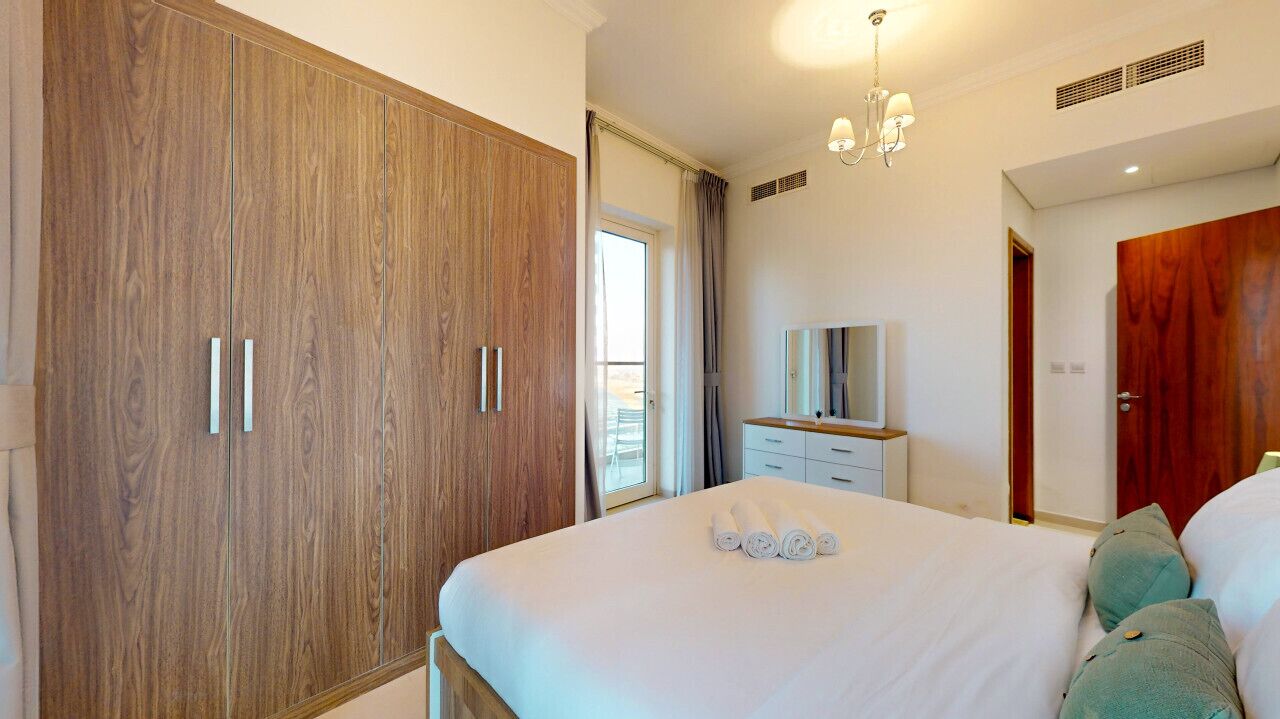 A bedroom with a large wardrobe, a bed with rolled towels, a bedside table, and a mirror.