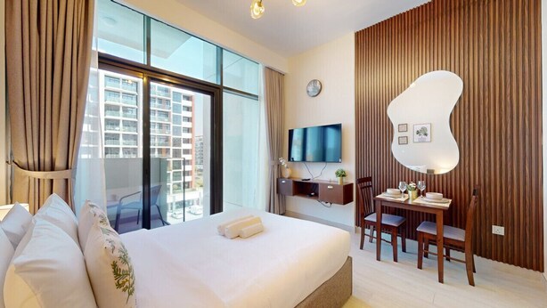 A hotel room with a large bed, a desk with a chair, a TV, and a view of the city through large windows. - Azizi Riviera 8 - Quiet Studio in Meydan (Meydan)