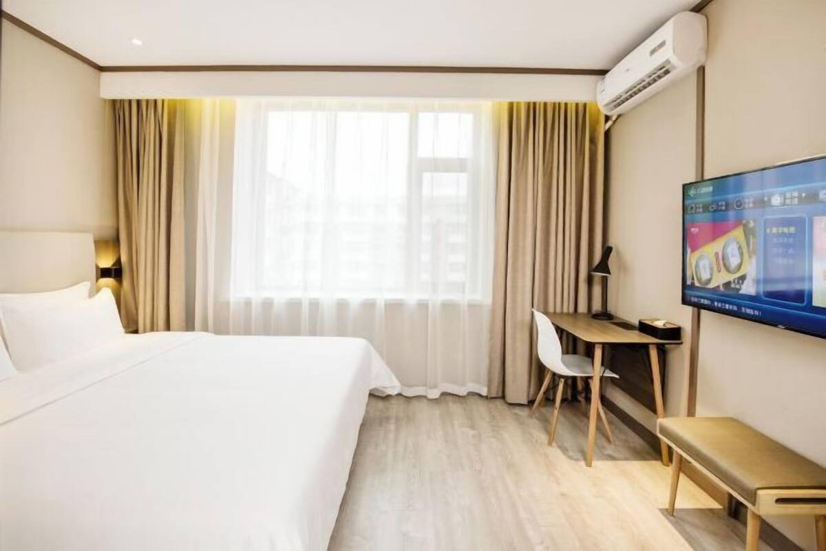 A hotel room with a bed, a desk, a chair, a TV, and an air conditioning unit.