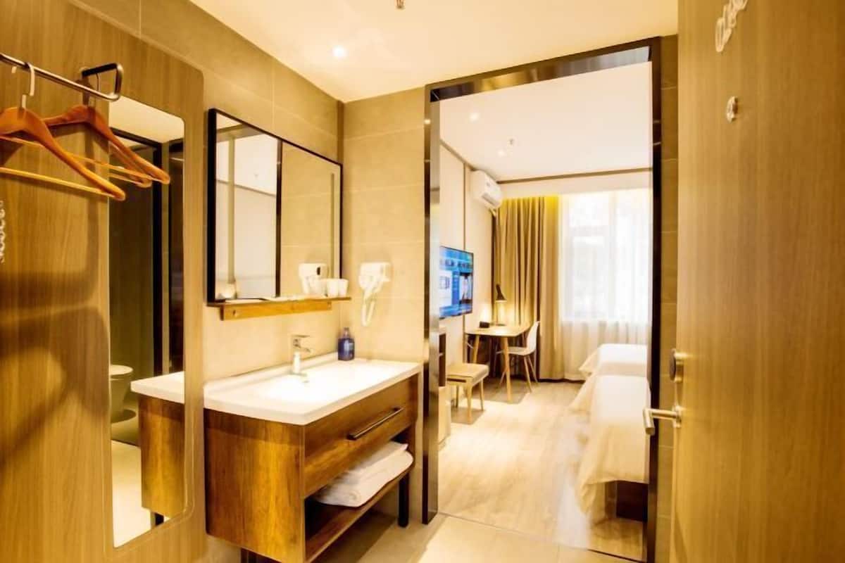 A hotel room with a wooden vanity, a mirror, a sink, a towel rack, a desk with a chair, and a bed.