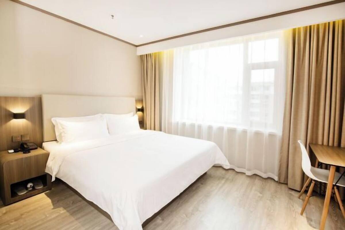 A hotel room with a large bed, bedside table with a lamp, a small table with a chair, and a window with curtains.