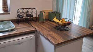 A compact kitchen with a wooden countertop, a wall clock, a fruit bowl, and a window with curtains.
