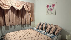 A bedroom with a bed, a window with curtains, a wall with a flower painting, and a bedside table with a lamp.