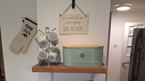 A kitchen with a wooden shelf holding mugs and a kettle, a hanging sign, and a green pot.