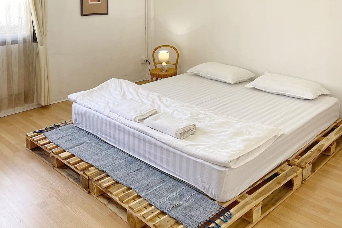 Une chambre avec un lit simple sur une plateforme en bois, une petite table de chevet avec une lampe, un tableau encadré au mur et une fenêtre avec des rideaux.