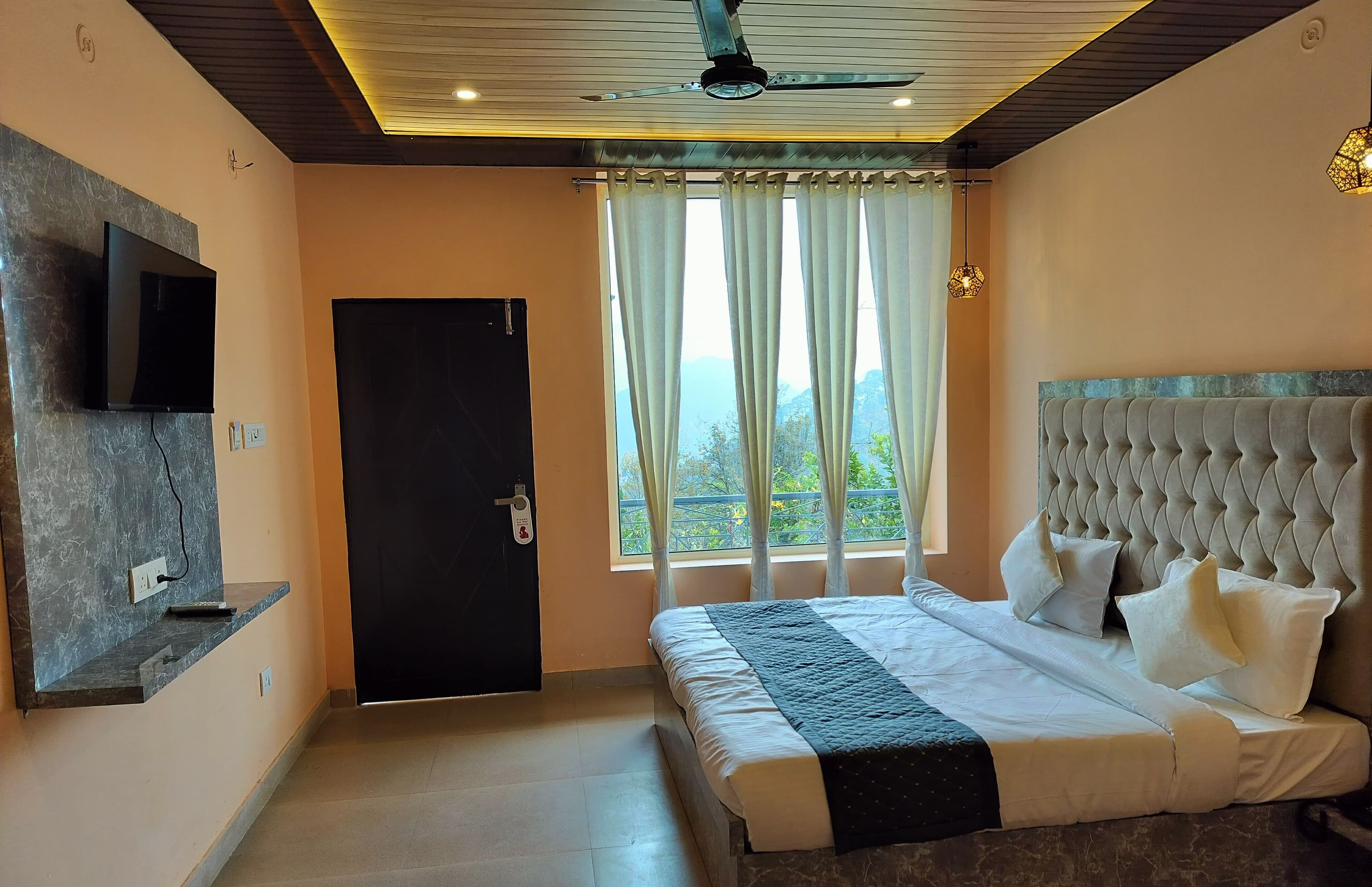 A bedroom with a bed, a TV mounted on the wall, a ceiling fan, and a window with curtains.