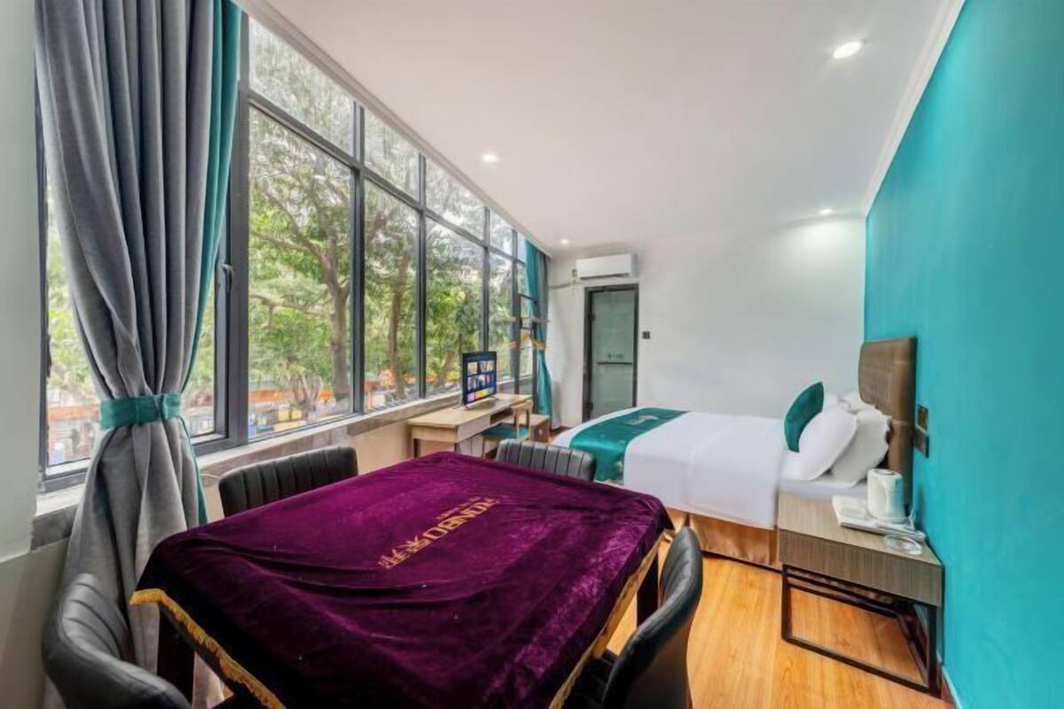 Une chambre d’hôtel avec une grande fenêtre, un lit recouvert d’une couverture verte, un bureau avec un ordinateur et une couverture violette sur la chaise.