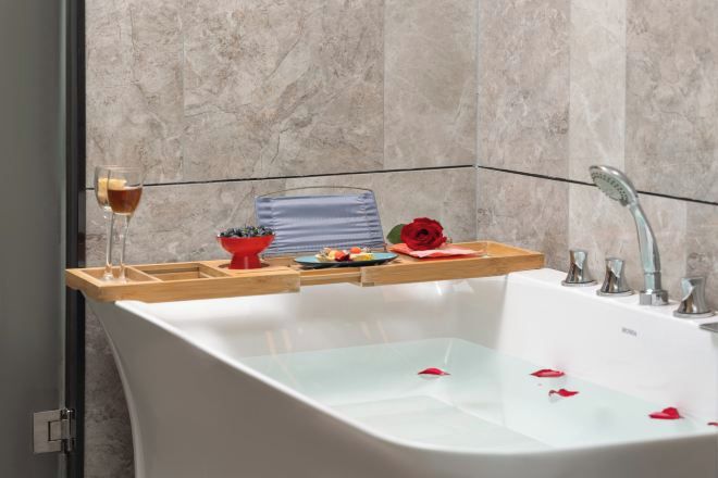 A modern bathroom with a freestanding bathtub, a glass of wine, a bowl of fruit, and a rose on the edge.