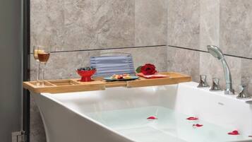 A modern bathroom with a freestanding bathtub, a glass of wine, a bowl of fruit, and a rose on the edge.