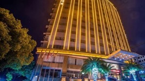 Un grand hôtel à la façade incurvée, illuminé par des jeux de lumière verticaux, et doté d’une entrée couverte agrémentée de palmiers.