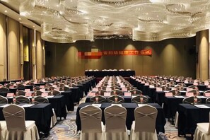 Une salle de conférence avec des rangées de tables et de chaises, une scène à l’avant et une grande bannière accrochée au mur.