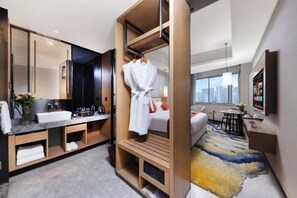 Une chambre d’hôtel moderne dotée d’un grand miroir, d’une coiffeuse intégrée, d’un peignoir à portée de main, d’un lit avec vue sur la ville et d’une petite table surmontée d’un vase.