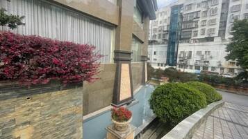 A residential building with a stone facade, a small garden area with a statue, and a view of a multi-story building in the background.