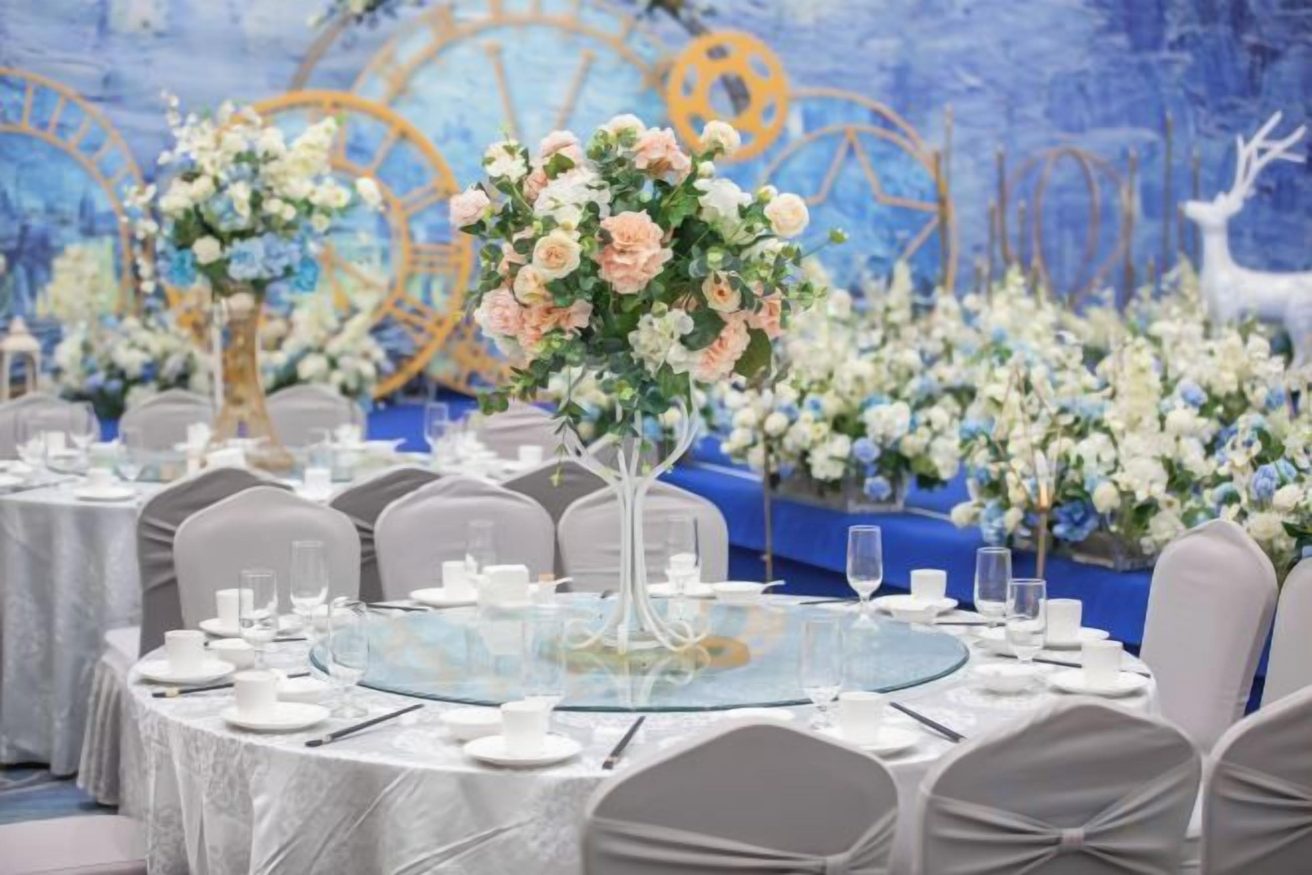Ein Banketthaus mit runden Tischen, festlich für eine Veranstaltung hergerichtet, mit floralen Tischdekorationen und einem blauen und goldenen Hintergrund.