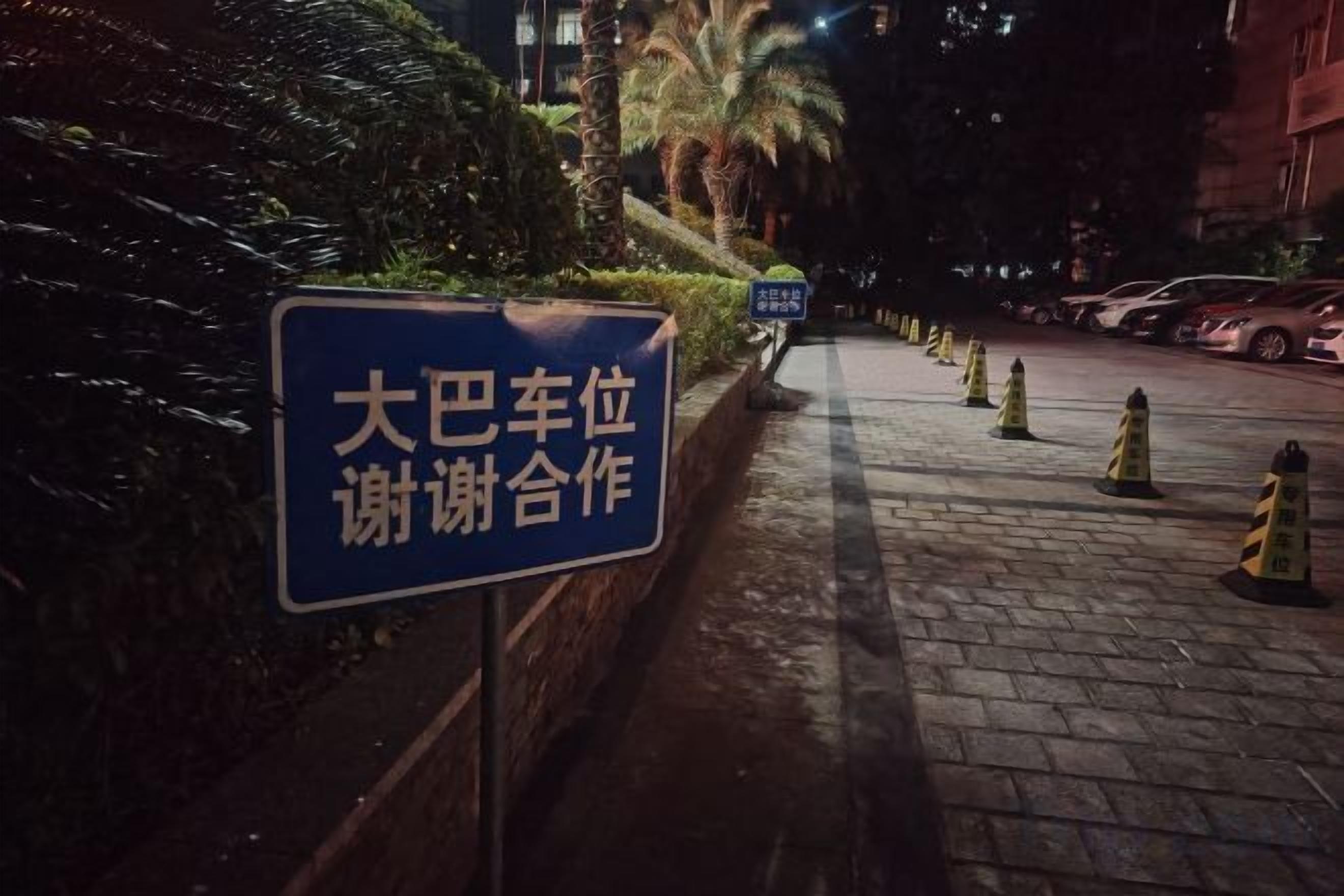 Scena notturna di una strada con un cartello blu sul marciapiede, pavimentazione in mattoni e coni stradali gialli.