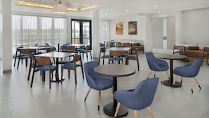 Un'area ristorazione moderna in un hotel, con tavoli rotondi e sedie blu, ampie finestre con tende e opere d'arte alle pareti.