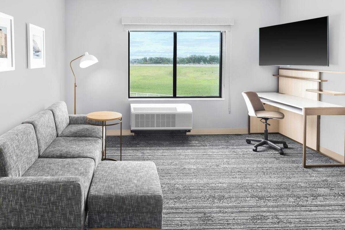Una habitación moderna con un sofá seccional gris, un escritorio con una silla, una lámpara de pie y un ventanal con vistas a un campo.