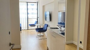 A modern hotel room with a wooden floor, a dining area with blue chairs, a TV mounted on the wall, and a large window with curtains. - orbi city apartments (Batumi)