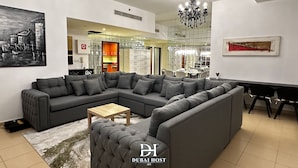 Ein modernes Wohnzimmer mit einem grauen Ecksofa, einem Holztisch und einer Kristallleuchtre. An der Wand hängt ein Gemälde, und im Hintergrund ist ein Essbereich zu sehen.