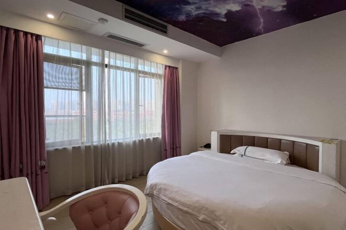 A hotel room with a bed, a chair, a desk, and a view of the city through large windows.