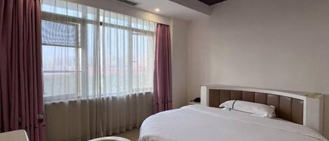 A hotel room with a bed, a chair, a desk, and a view of the city through large windows.
