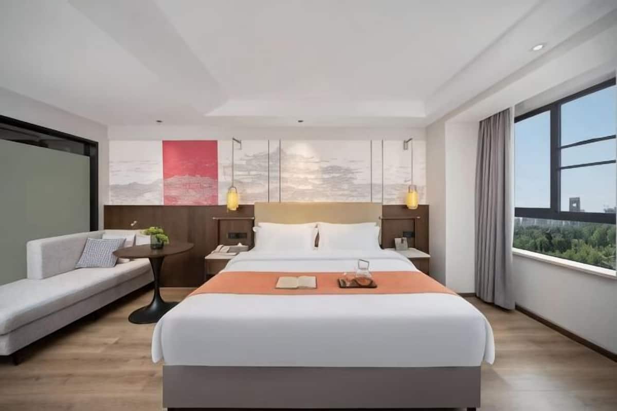 A modern hotel room with a large bed, a sofa, a nightstand, a lamp, and a view of the city.