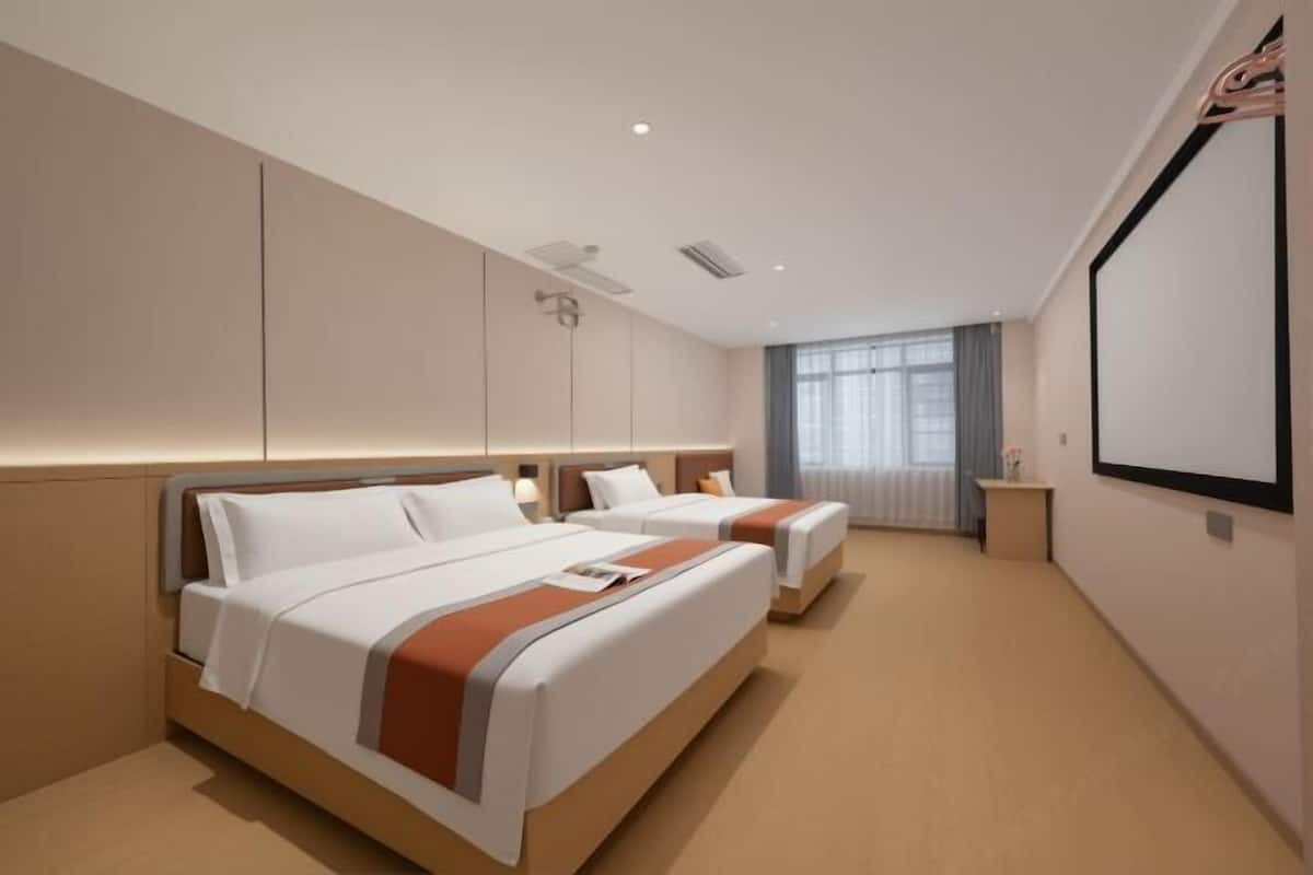 A hotel room with two beds, a large window with curtains, a flat-screen TV mounted on the wall, and a small bedside table.