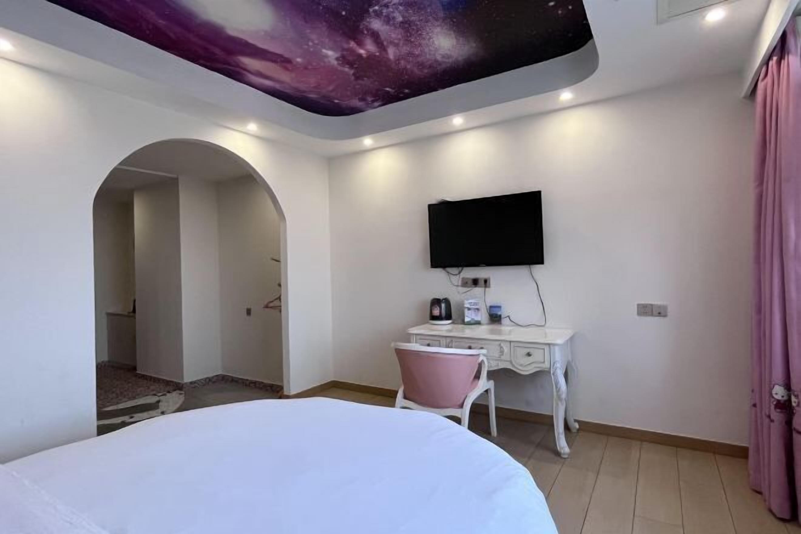 Una habitación de hotel con cama, un escritorio con silla, un televisor de pantalla plana y un mural en el techo.